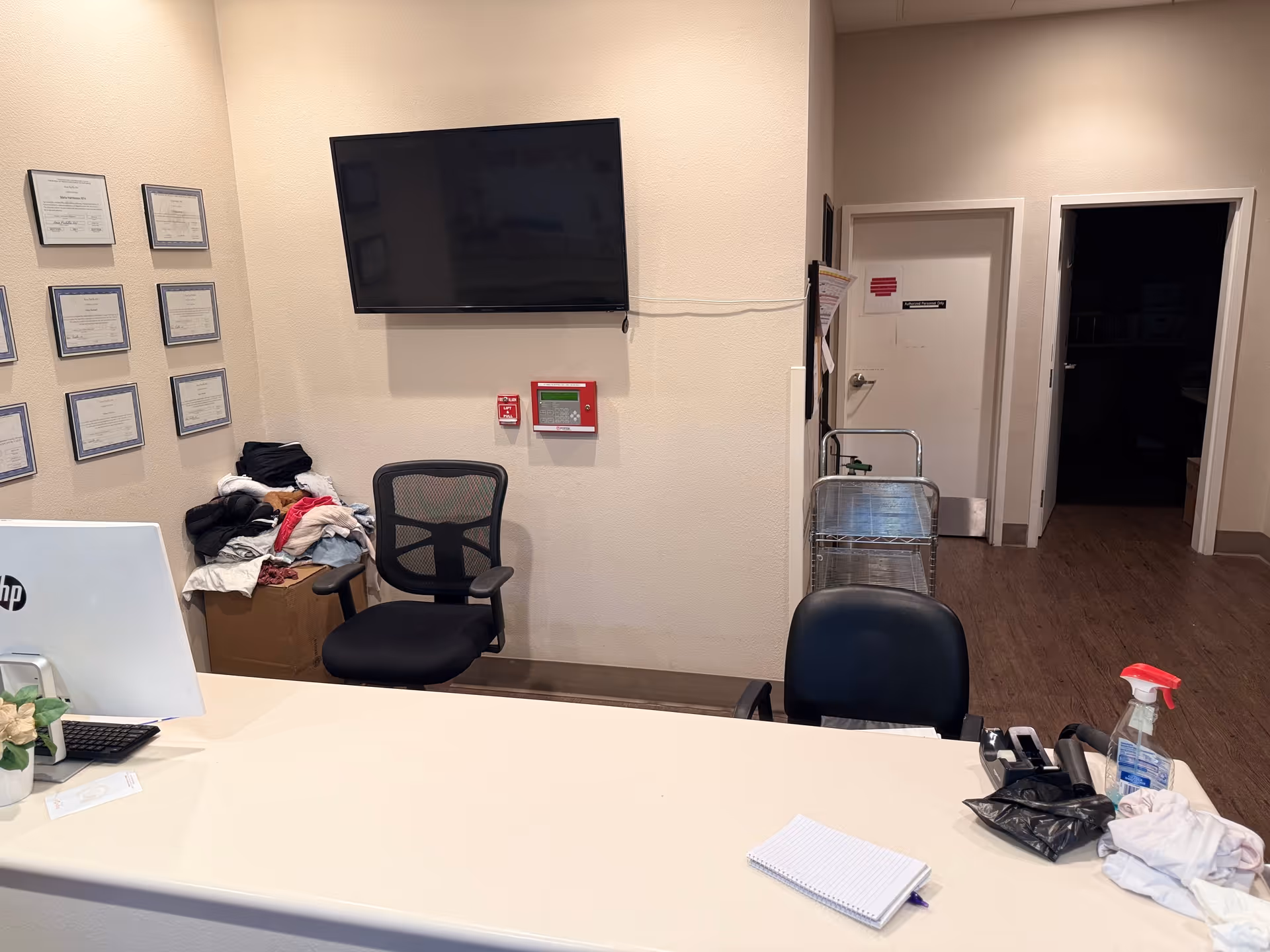Reception desk and small office area with chairs, a wall-mounted TV, certificates on the wall, a cart, and cleaning supplies.