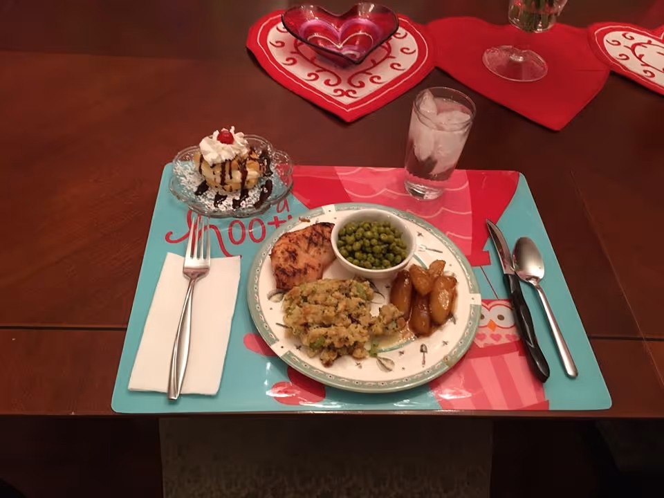 A meal served on a colorful placemat on a wooden dining table. The plate contains cooked green peas in a small bowl, roasted potatoes, stuffing, and a piece of grilled chicken. To the left of the plate is a fork on a white napkin, and to the right are a knife and spoon. Above the plate is a glass of water with ice and a dessert dish with a small cake topped with whipped cream and a cherry. The table has red heart-shaped placemats and a glass vase with flowers in the background.
