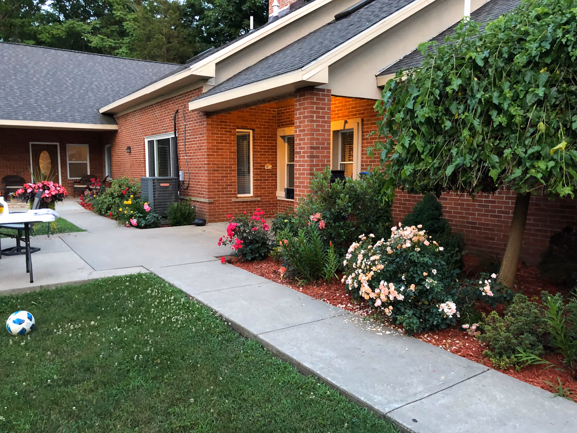 Brick single-story building entrance and courtyard with a sidewalk, flowering shrubs, patio table and a grassy lawn.