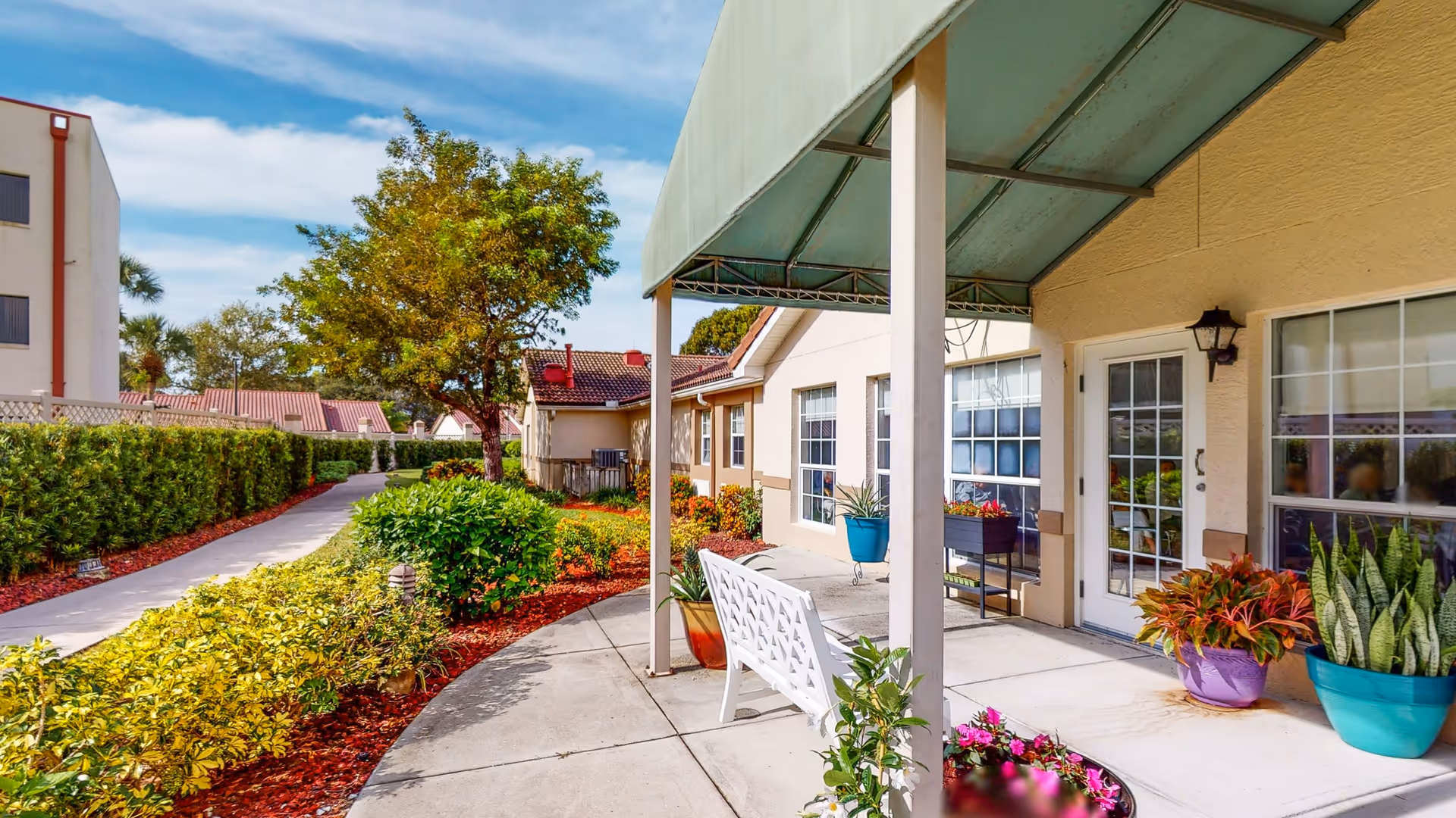 Outdoor patio area of a senior living community with a white bench, potted plants, and a covered awning. A concrete pathway curves through landscaped greenery and bushes, leading to other buildings under a partly cloudy blue sky.