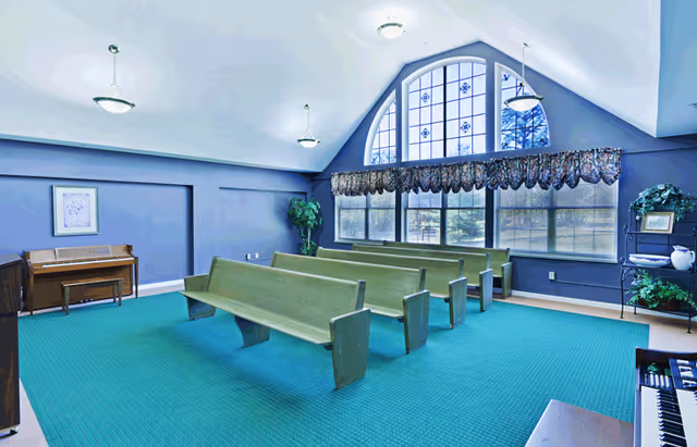 Bright interior room with green wooden pew-style benches facing a large arched window and a piano.
