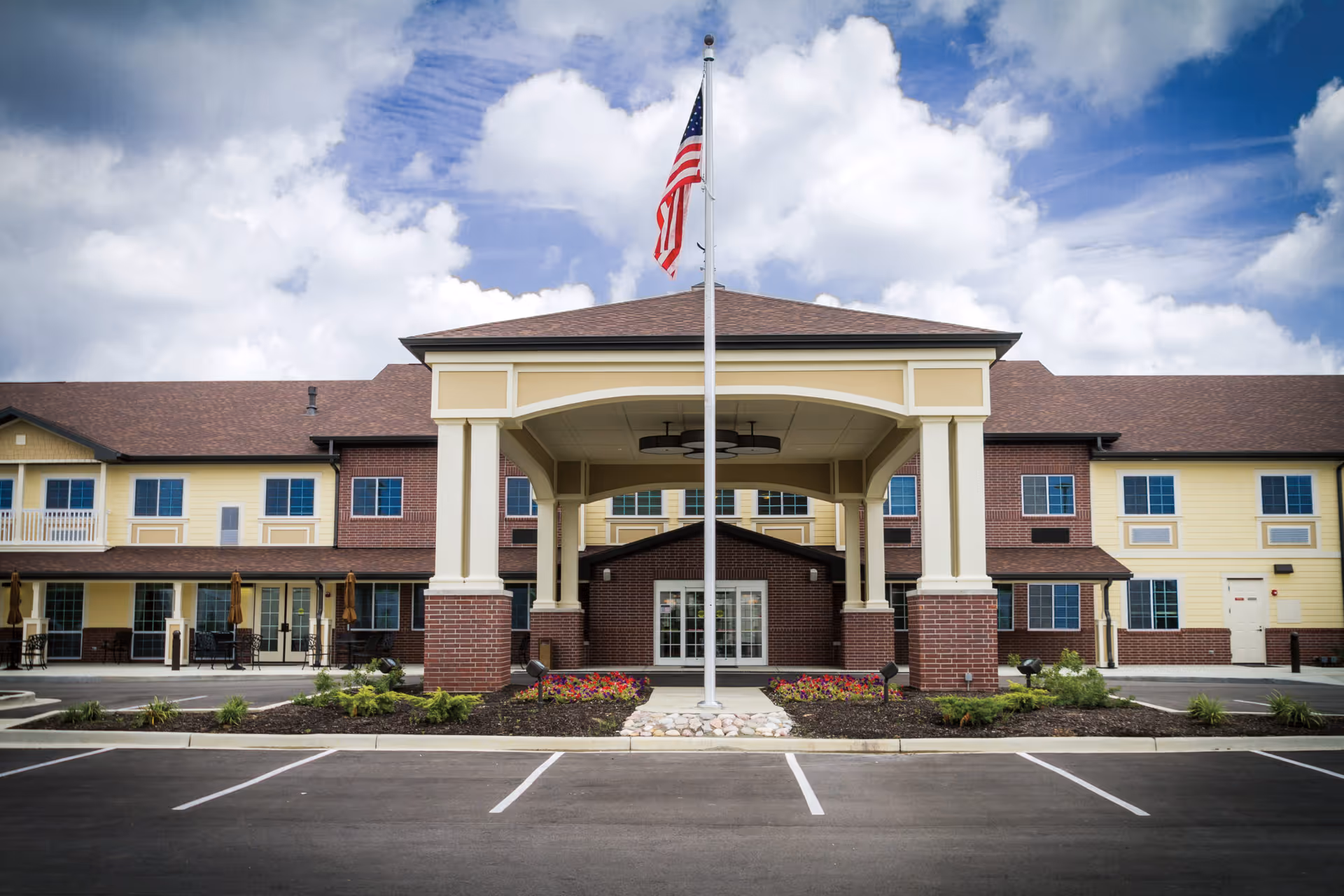 Entrance of a two-story senior living building with a covered portico, American flag on a pole, landscaping, and empty parking spaces.
