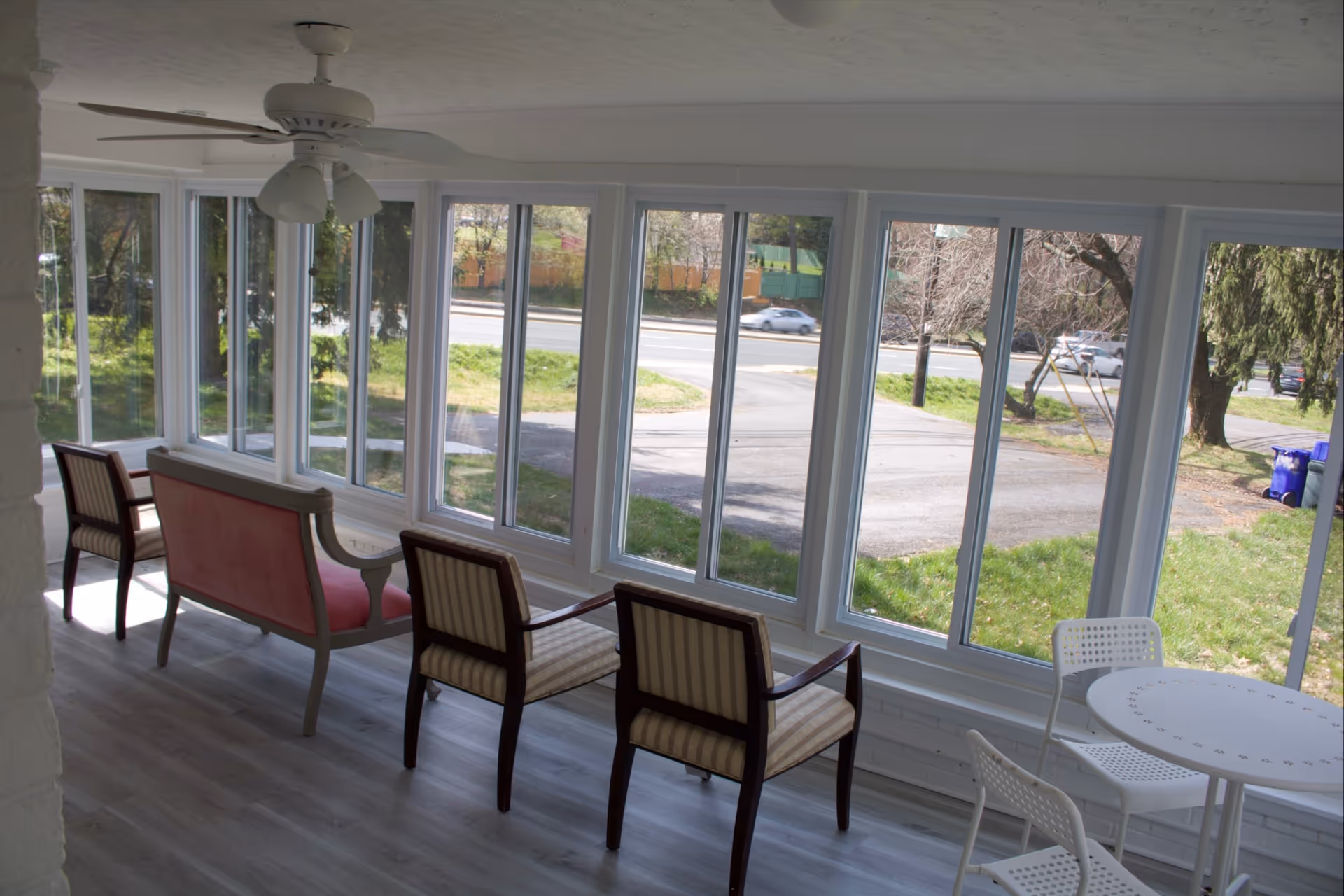A bright sunroom with large windows overlooking a grassy outdoor area and a road. The room contains a ceiling fan, two striped armchairs, a pink cushioned loveseat, and a small white round table with two white chairs.