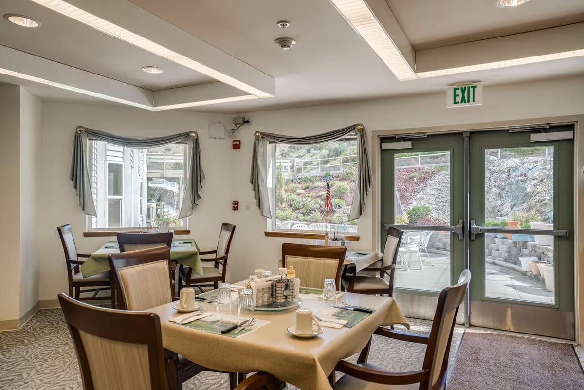 A dining area in a senior living facility with tables covered in beige and green tablecloths, set with cups, glasses, and utensils. The room has large windows with draped curtains and a glass door leading to an outdoor patio with chairs and a landscaped garden.