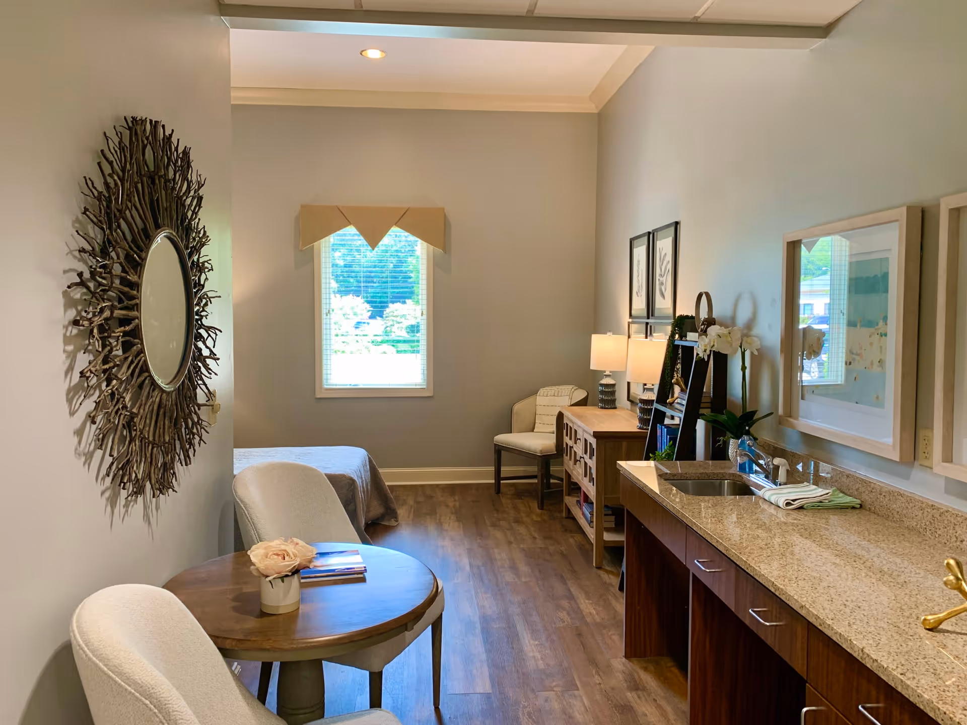 A cozy interior room with light gray walls and wooden flooring. On the left, there is a round wooden table with two white upholstered chairs and a small flower vase on the table. A decorative circular mirror with a natural twig frame hangs on the wall. In the background, a window with blinds lets in natural light. On the right side, there is a granite countertop with a sink, a wooden cabinet, a small bookshelf with books and decor, a chair, and two table lamps. Framed artwork hangs on the walls.