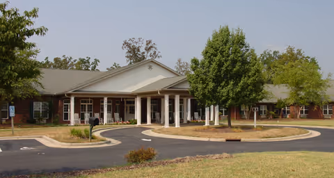 Front entrance of a single-story brick senior living facility with a covered porte-cochere and circular driveway surrounded by trees and lawn.