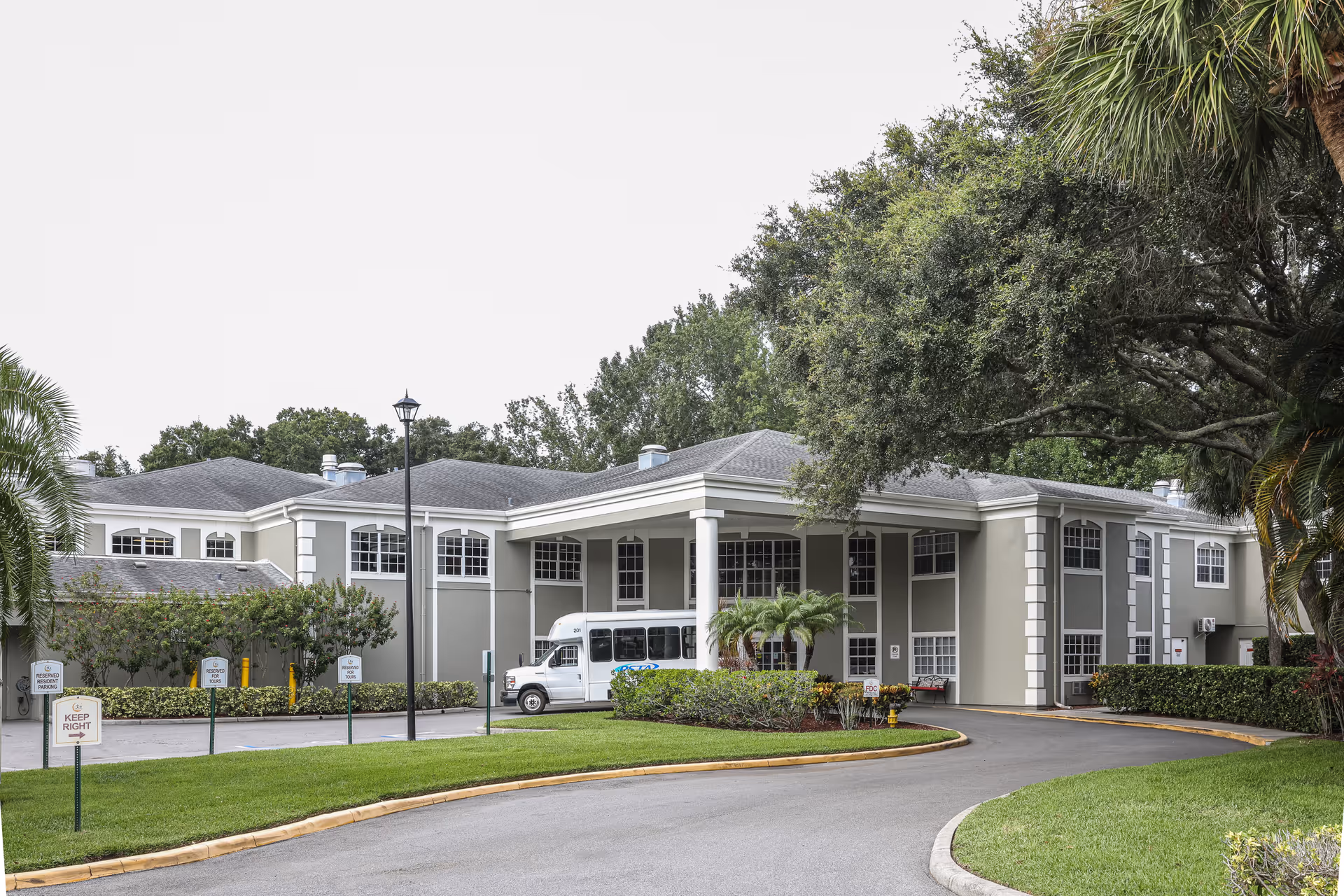 Exterior view of The Gables of Palm Harbor senior living facility showing a two-story building with a covered entrance, surrounded by trees and greenery, with a driveway and a white shuttle bus parked near the entrance.
