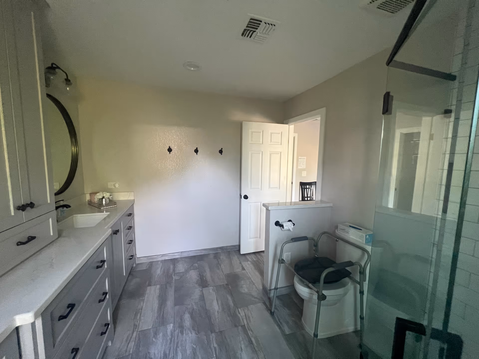 A spacious bathroom with gray tiled floor and light gray walls. On the left side, there is a long vanity with multiple drawers, a white countertop, a sink, and a round mirror above it. On the right side, there is a toilet equipped with a safety frame and a box of tissues on a small half-wall next to it. A glass-enclosed shower is partially visible on the far right. The bathroom door is open, showing a glimpse of an adjacent room with a chair.