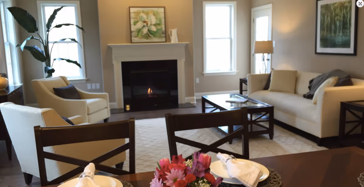 A cozy living room with a beige sofa, two beige armchairs, a dark wood coffee table, and a fireplace with a floral painting above it. There are two windows letting in natural light, a floor lamp, and a dining table in the foreground set with plates, napkins, and a colorful flower centerpiece.