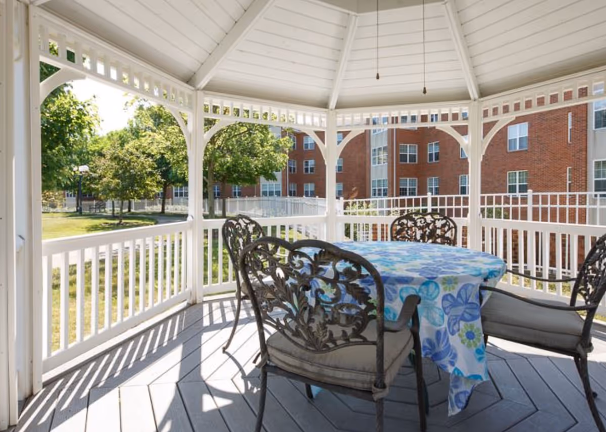 White gazebo with a round table covered by a blue floral tablecloth and four metal chairs overlooking a grassy courtyard and brick apartment building.