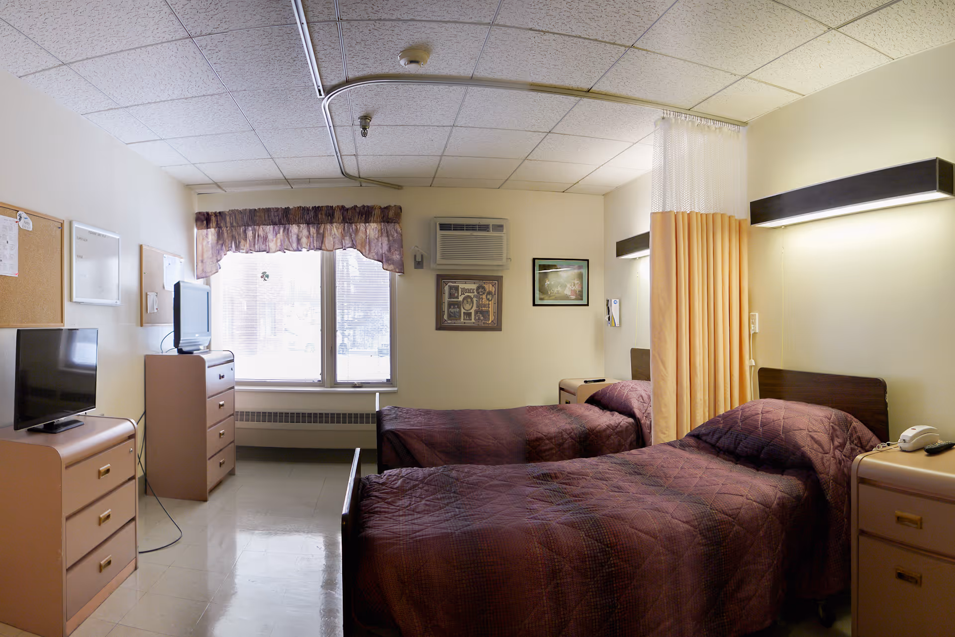 A shared bedroom in a senior living facility with two single beds separated by a privacy curtain. Each bed has a dark purple quilted bedspread and a bedside table with a telephone. The room has a window with blinds and a valance, a wall-mounted air conditioning unit, two dressers with televisions on top, and framed pictures on the wall.