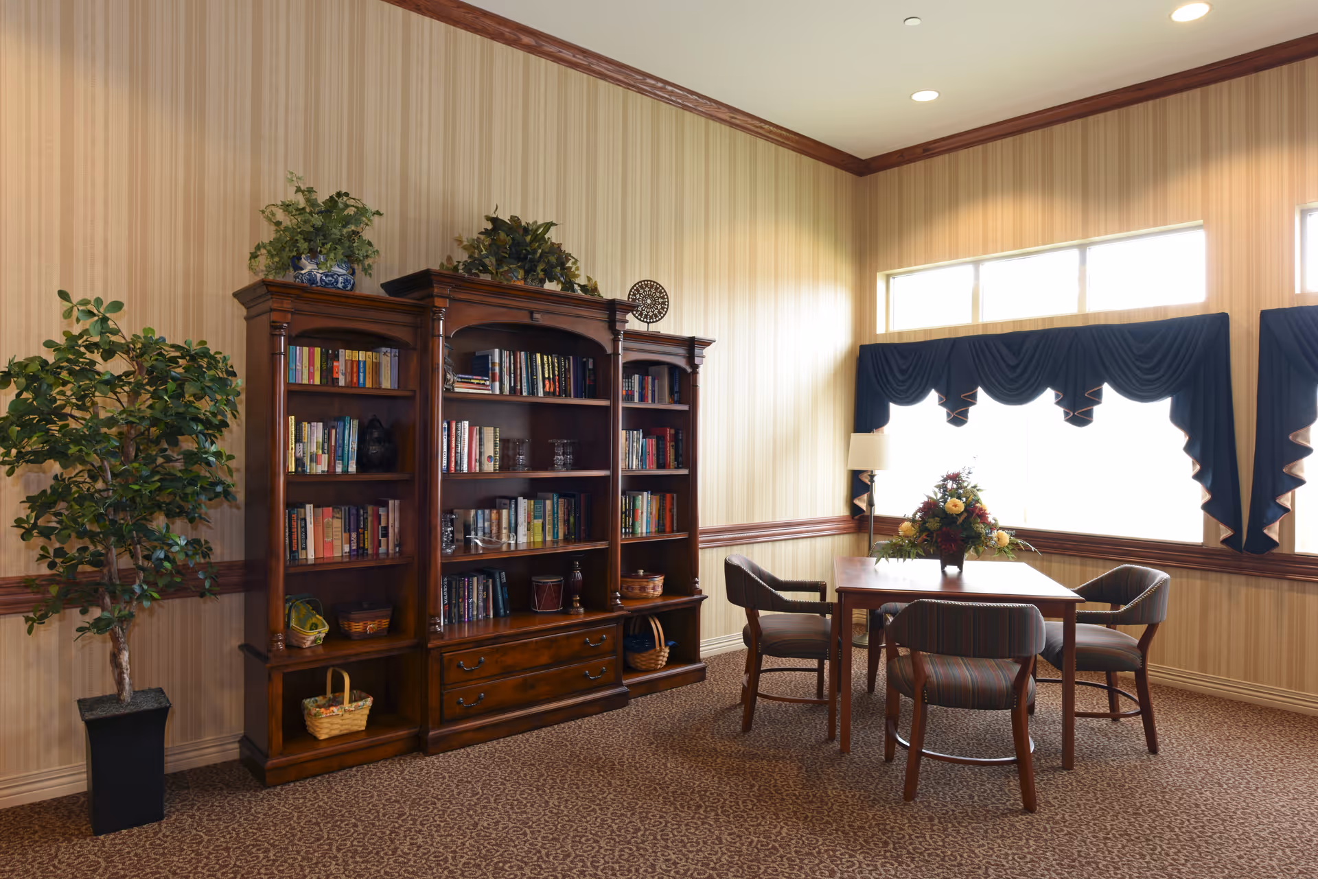 A cozy interior room with a wooden bookshelf filled with books and decorative items, a potted plant to the left, and a wooden table with four upholstered chairs around it. The room has beige striped wallpaper, carpeted floor, and large windows with dark blue valances letting in natural light. A floral arrangement is placed on the table.