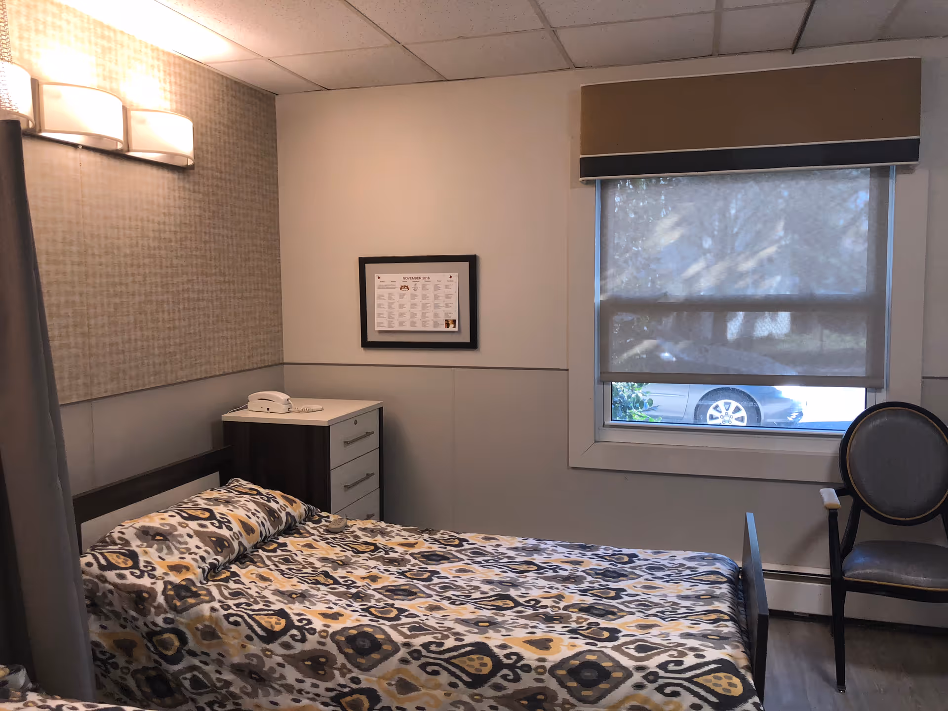 Small furnished bedroom with a single bed, nightstand, chair, and a window with a roller shade.