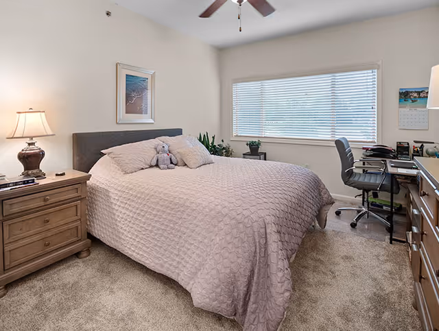 A bedroom with a large bed covered in a textured beige bedspread, two pillows, and a small stuffed animal. There is a wooden nightstand with a lamp and some items on it to the left of the bed. A large window with white blinds is on the right wall, next to a desk with an office chair and various office supplies. The room has beige carpet and light-colored walls with a framed picture above the bed.