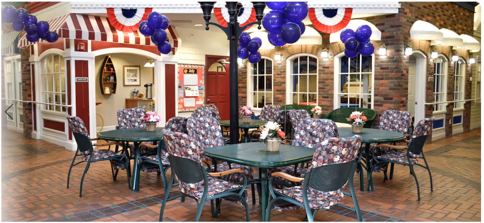 Indoor seating area with multiple round tables and floral cushioned chairs arranged around them. Each table has a small flower arrangement. The space is decorated with purple balloons and red, white, and blue bunting. The background features a brick wall with windows and a small reception or office area with a red door and bulletin board.