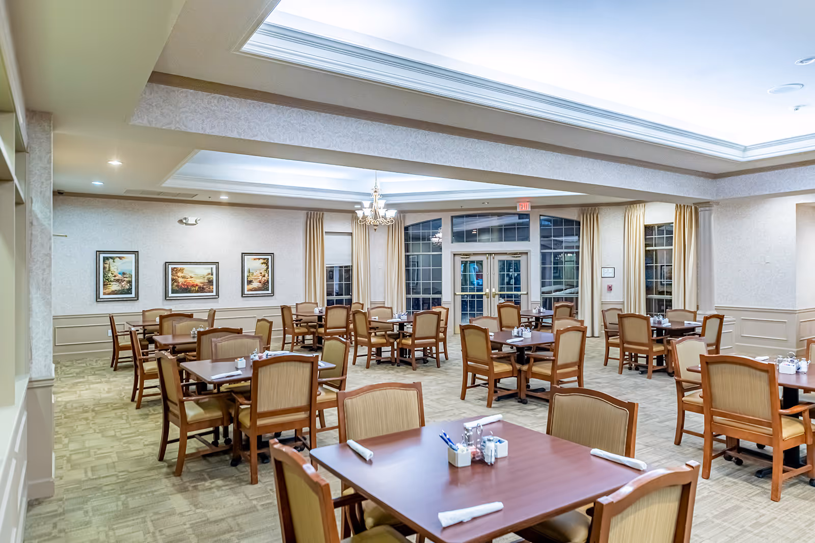 A spacious dining room with multiple wooden tables and cushioned chairs arranged neatly. The room has large windows with beige curtains, soft lighting from ceiling fixtures, and framed landscape paintings on the walls. Each table is set with napkins and small condiment holders.