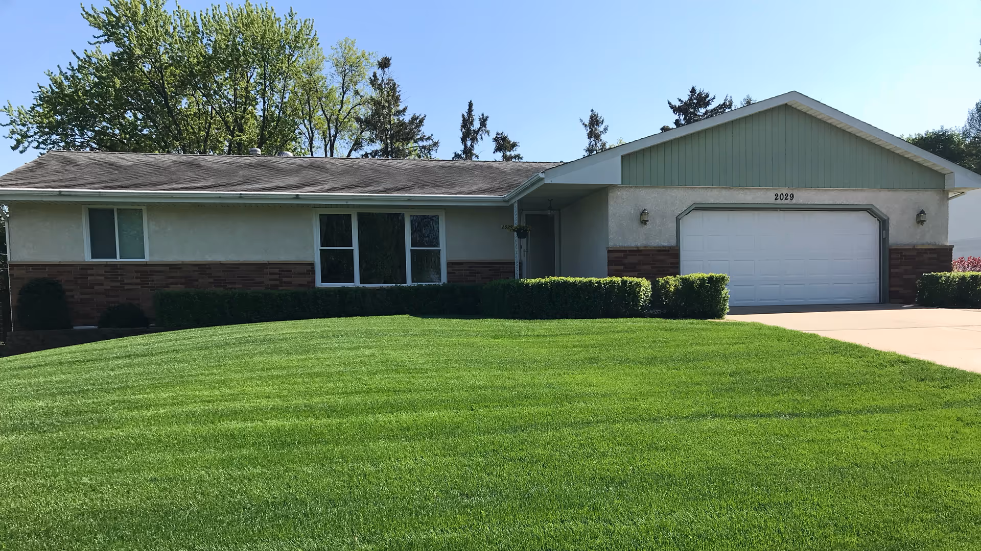 Single-story ranch-style house front with a large green lawn, attached two-car garage, and trimmed shrubs.