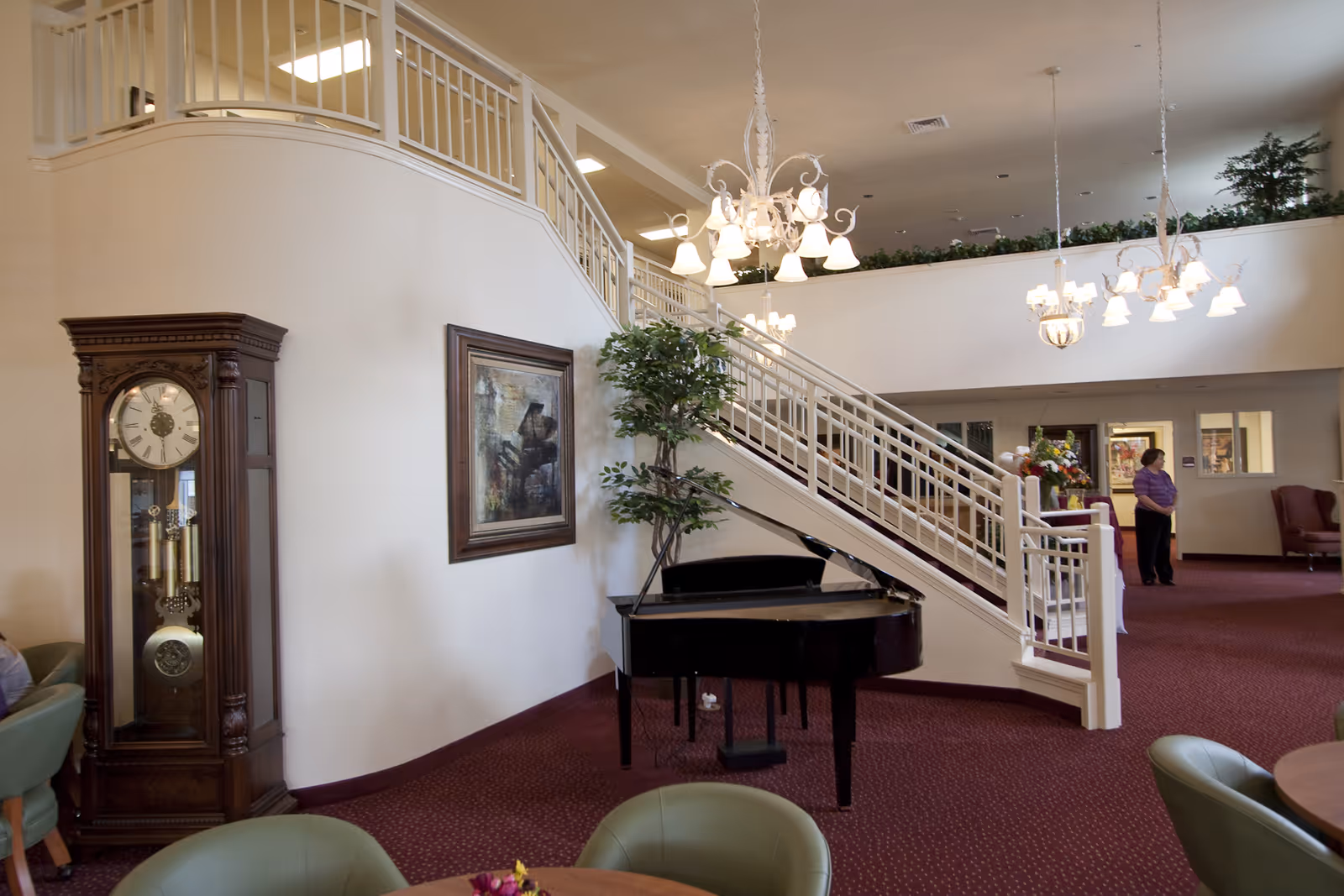 Interior view of a senior living facility lobby area featuring a grand piano, a tall grandfather clock, a framed painting, a staircase with white railings, green chairs around a wooden table, and a woman standing near the back. The space is carpeted in red with white walls and decorated with hanging chandeliers and plants.