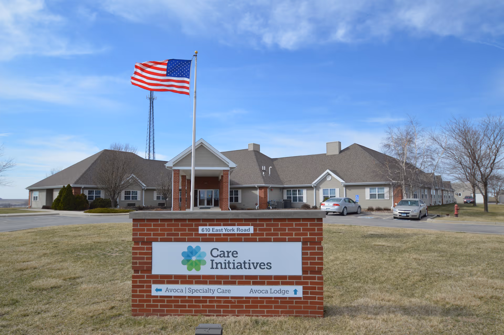 Exterior view of a single-story senior living facility building with a brick sign in front displaying 'Care Initiatives' and directional information for Avoca Specialty Care and Avoca Lodge. An American flag flies on a flagpole above the sign, and there are a few cars parked near the building under a partly cloudy sky.