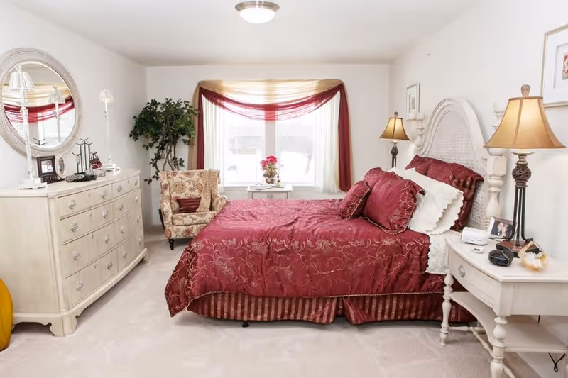 A cozy bedroom with a large bed covered in a red patterned comforter and matching pillows. The room features a white wooden headboard, two bedside tables with lamps, a white dresser with a round mirror above it, a floral upholstered armchair, a small white table with a flower vase, and a window with sheer curtains and a red swag valance. The carpet is light-colored and the walls are painted white.