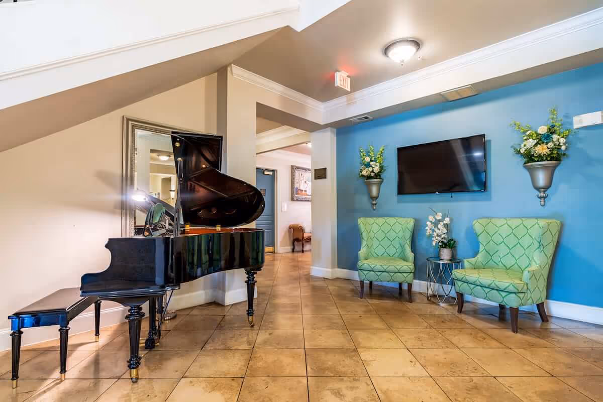 Lobby/common room with a black grand piano, two green armchairs, a wall-mounted TV and floral decor.