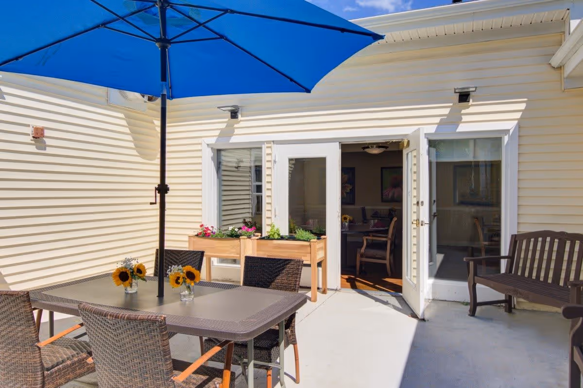 Outdoor patio area with a table and four wicker chairs under a large blue umbrella. There are small vases with sunflowers on the table. Two wooden planter boxes with flowers are placed near a set of white double doors leading inside. A wooden bench is positioned to the right of the doors. The building exterior is light yellow siding.