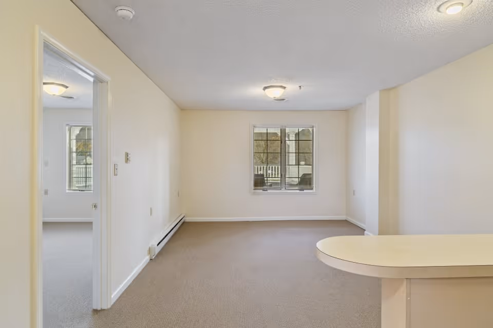 Empty open living room with beige walls and carpet, a center window, and a rounded kitchen counter visible in the foreground.