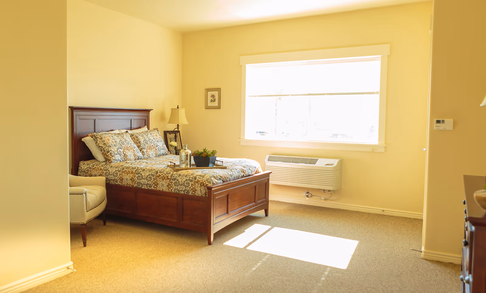 Sunny furnished bedroom with a wooden bed, patterned bedding, an armchair, nightstand with a lamp, and a large window.