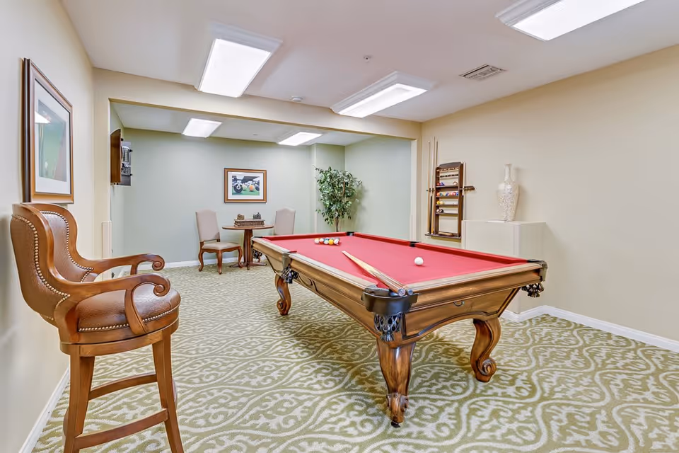 A game room with a red felt pool table in the center, two wooden chairs with brown cushions on the left, a small round table with two chairs in the back, a wall-mounted pool cue rack with cues, a decorative vase on a pedestal, a framed picture on the wall, and a patterned carpeted floor.
