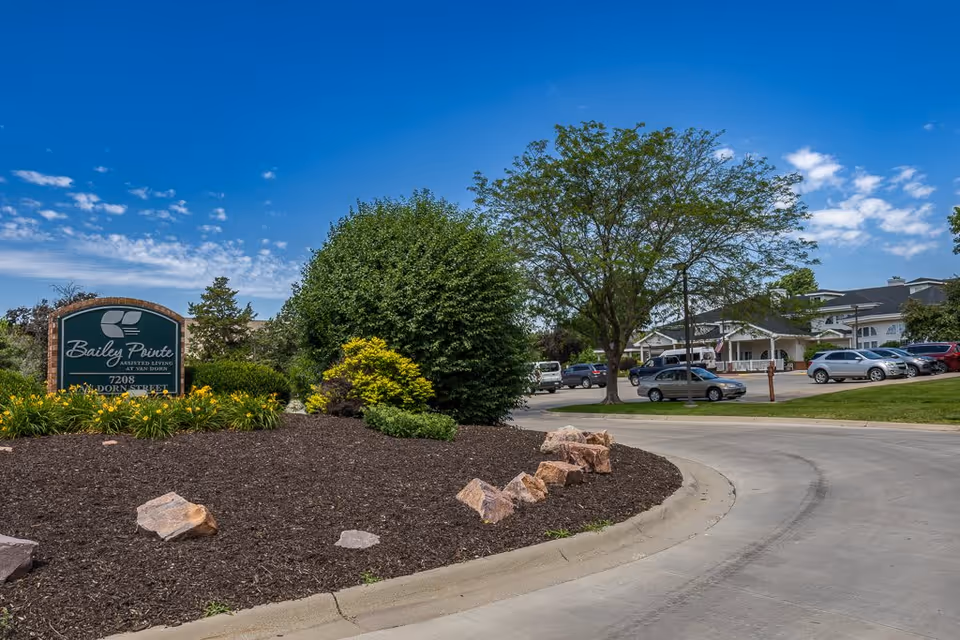 Exterior view of Bailey Pointe Assisted Living at Van Dorn showing the facility's entrance sign surrounded by landscaping with bushes and flowers, a curved driveway, parked cars, and a large tree under a blue sky with scattered clouds.