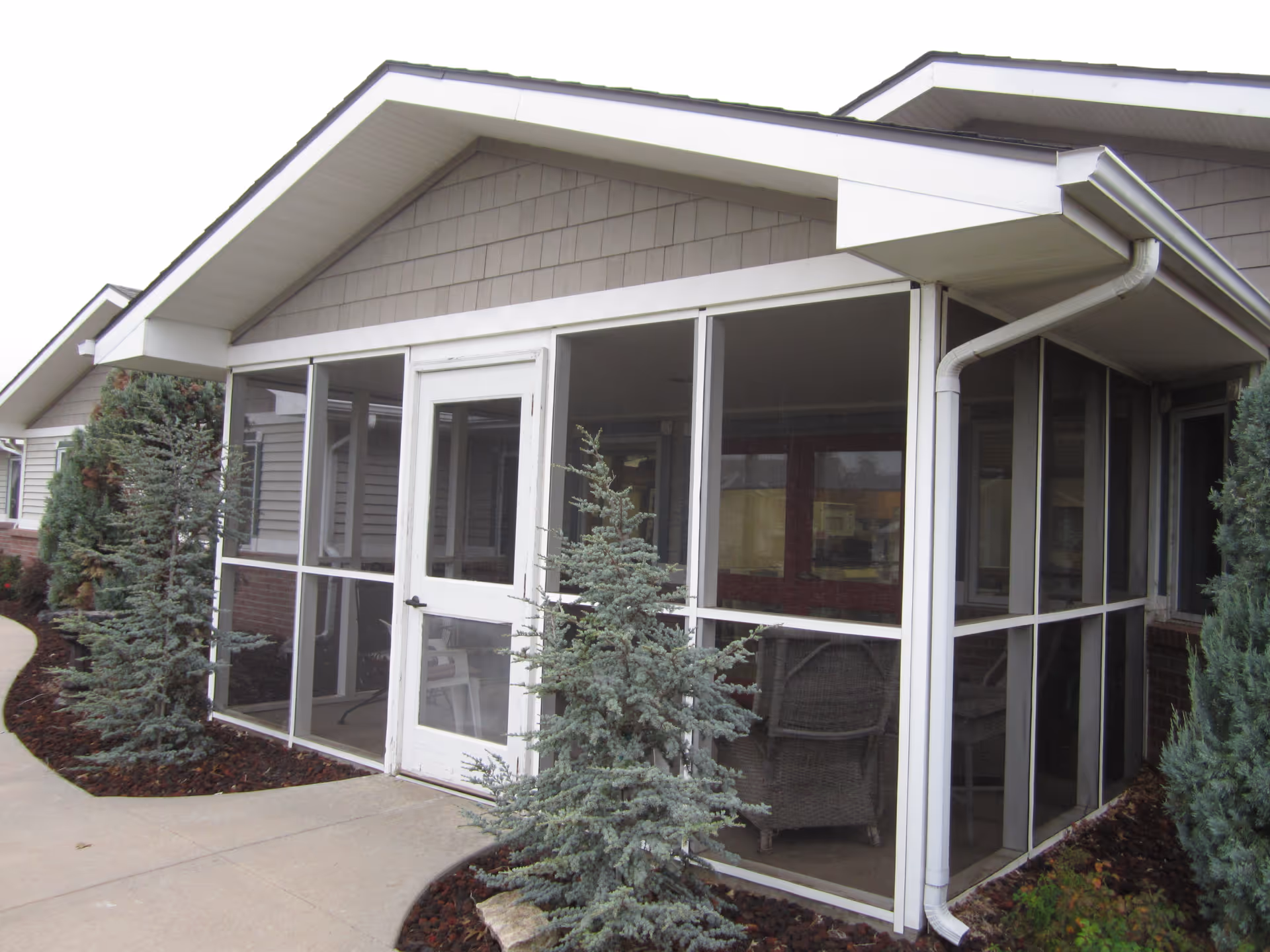 Exterior view of a screened-in porch attached to a building, with a white door and large screened windows. There are small evergreen trees and a curved concrete walkway in front of the porch.
