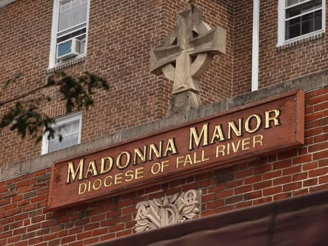 Brick building facade with a wooden sign reading "Madonna Manor Diocese of Fall River" beneath a stone cross.