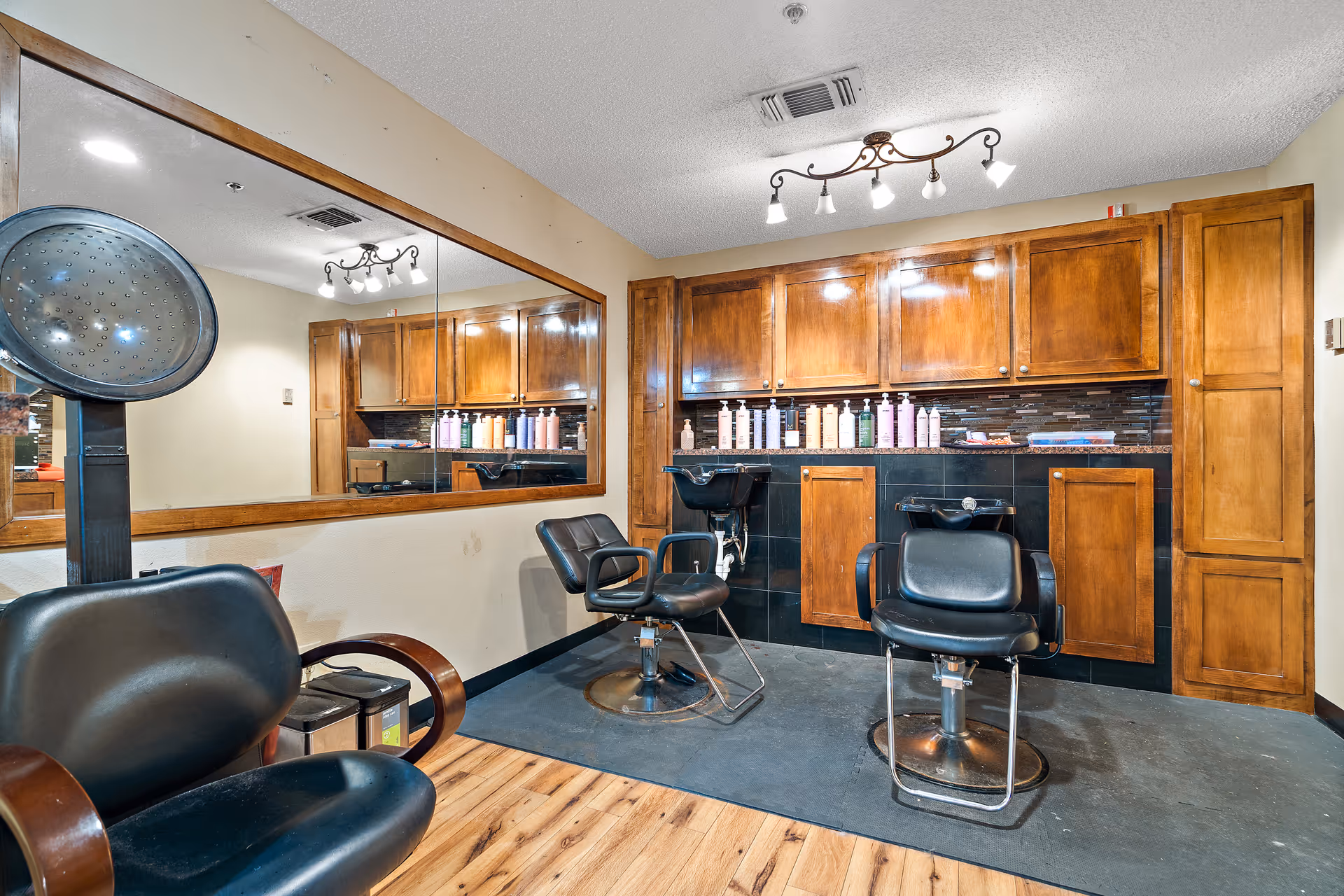 Interior view of a hair salon area in a senior living facility with two black salon chairs in front of wash basins, wooden cabinets above the basins, a large mirror on the wall, and a hair dryer chair in the foreground.