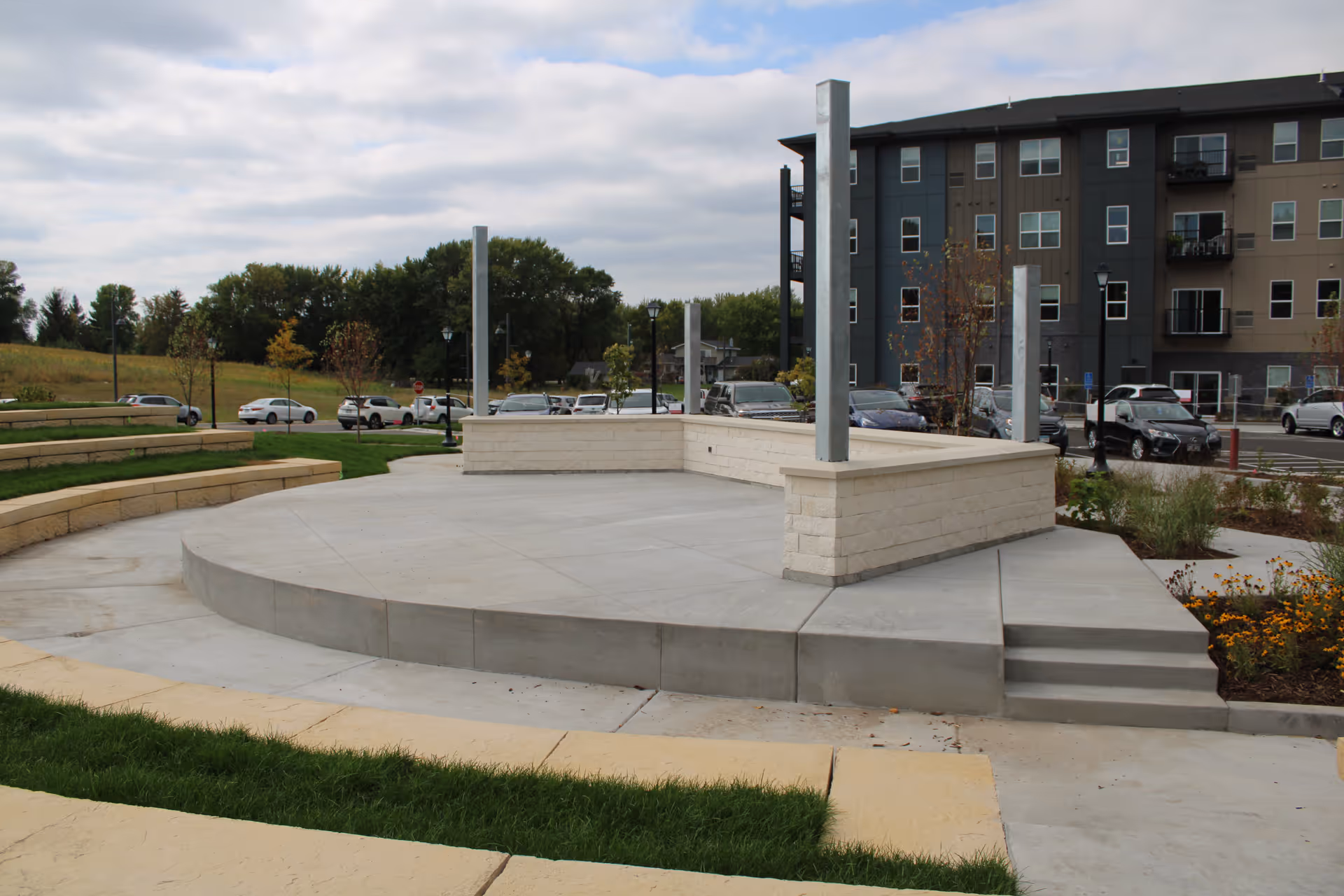 Outdoor amphitheater with curved concrete seating and a raised concrete stage area with metal posts. In the background, there is a parking lot with cars and a multi-story residential building. Trees and grassy areas surround the amphitheater under a cloudy sky.