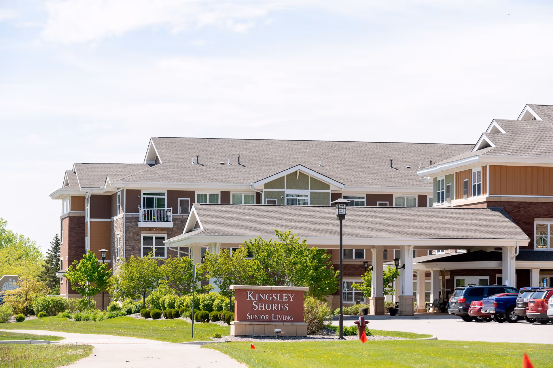 Exterior view of Kingsley Shores Senior Living facility showing a multi-story building with a covered entrance, several parked cars, green trees, and a sign in front that reads 'Kingsley Shores Senior Living'.