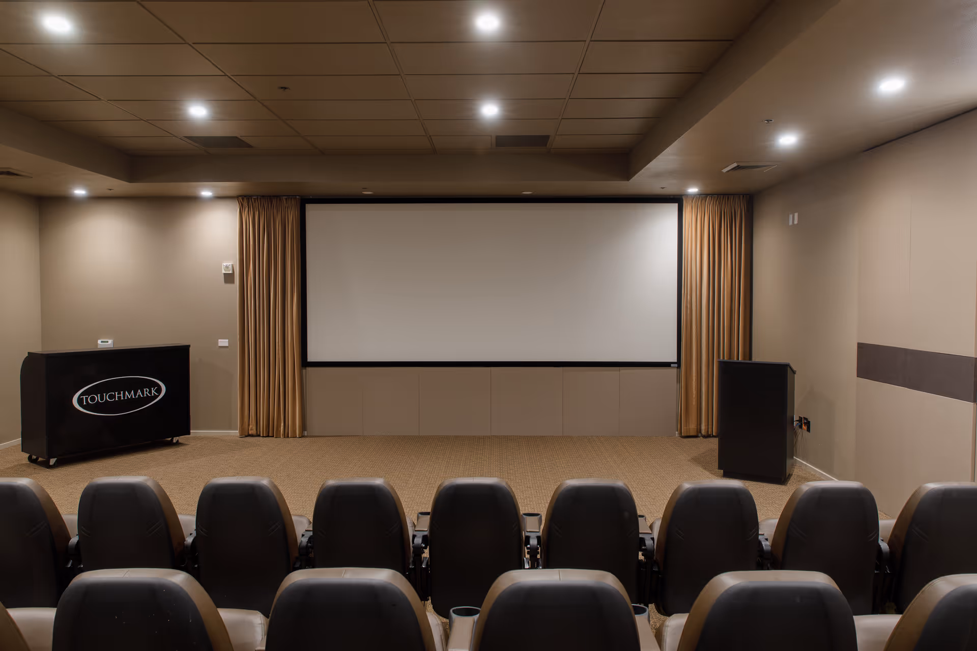 Interior view of a small theater or presentation room with rows of black cushioned seats facing a large blank projection screen. The room has beige walls, a carpeted floor, ceiling lights, and two podiums on either side of the screen. One podium has the word 'TOUCHMARK' on it.