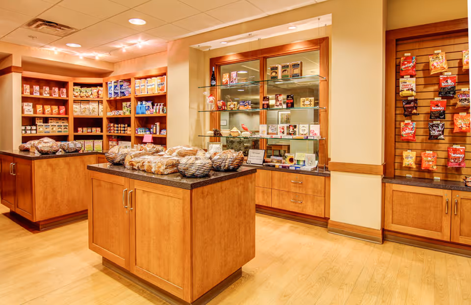 Interior view of a small convenience store or snack shop with wooden shelves and cabinets. The shelves are stocked with various packaged snacks, bread, and other grocery items. There are baskets of bread on two central wooden islands, and the lighting is warm and inviting.