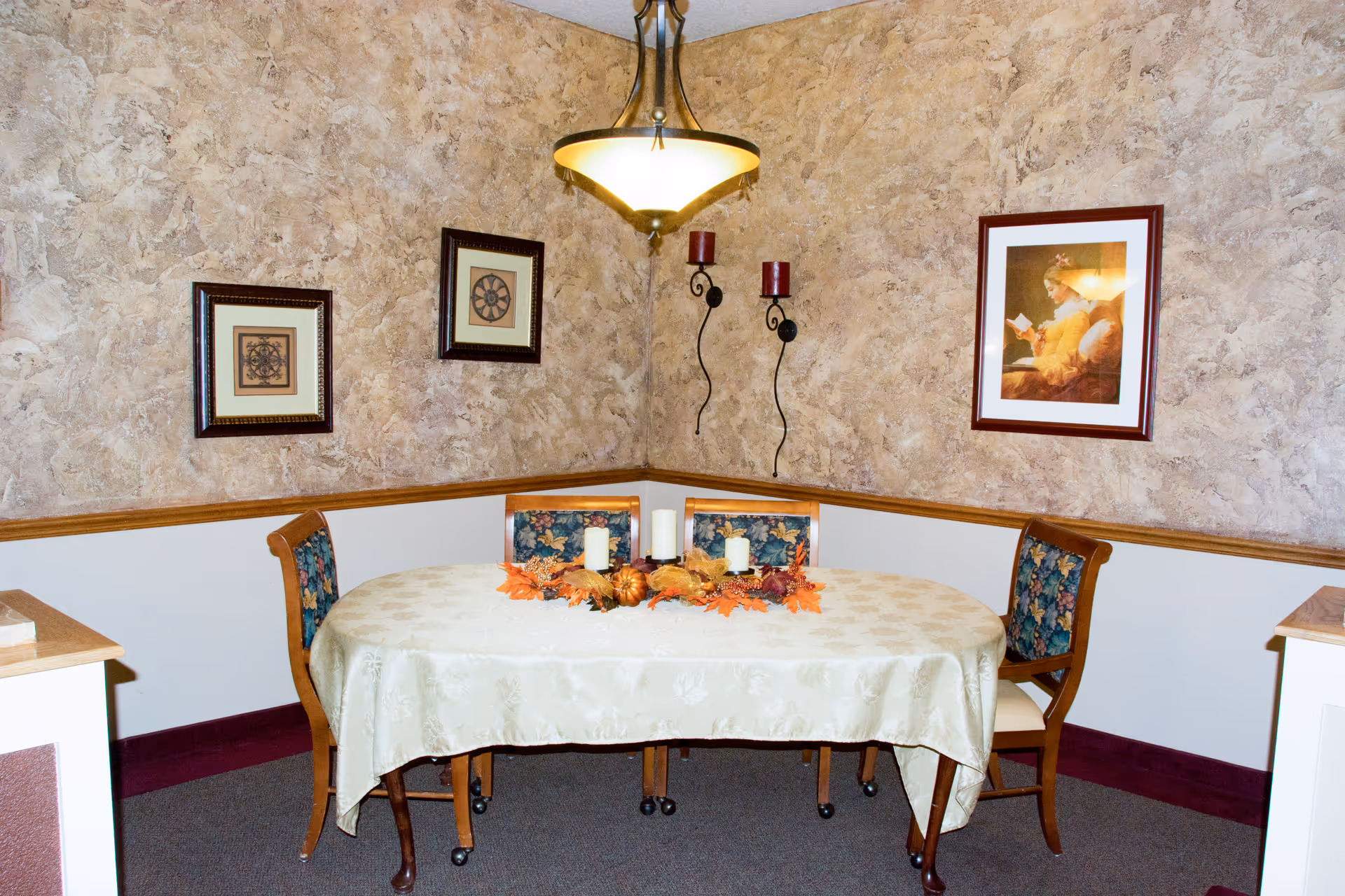 A small dining area with an oval table covered by a cream-colored tablecloth. The table is decorated with a centerpiece featuring autumn leaves and candles. Four wooden chairs with floral upholstery surround the table. The walls have a textured beige finish with three framed pictures and two wall-mounted candle holders. A hanging light fixture illuminates the space.