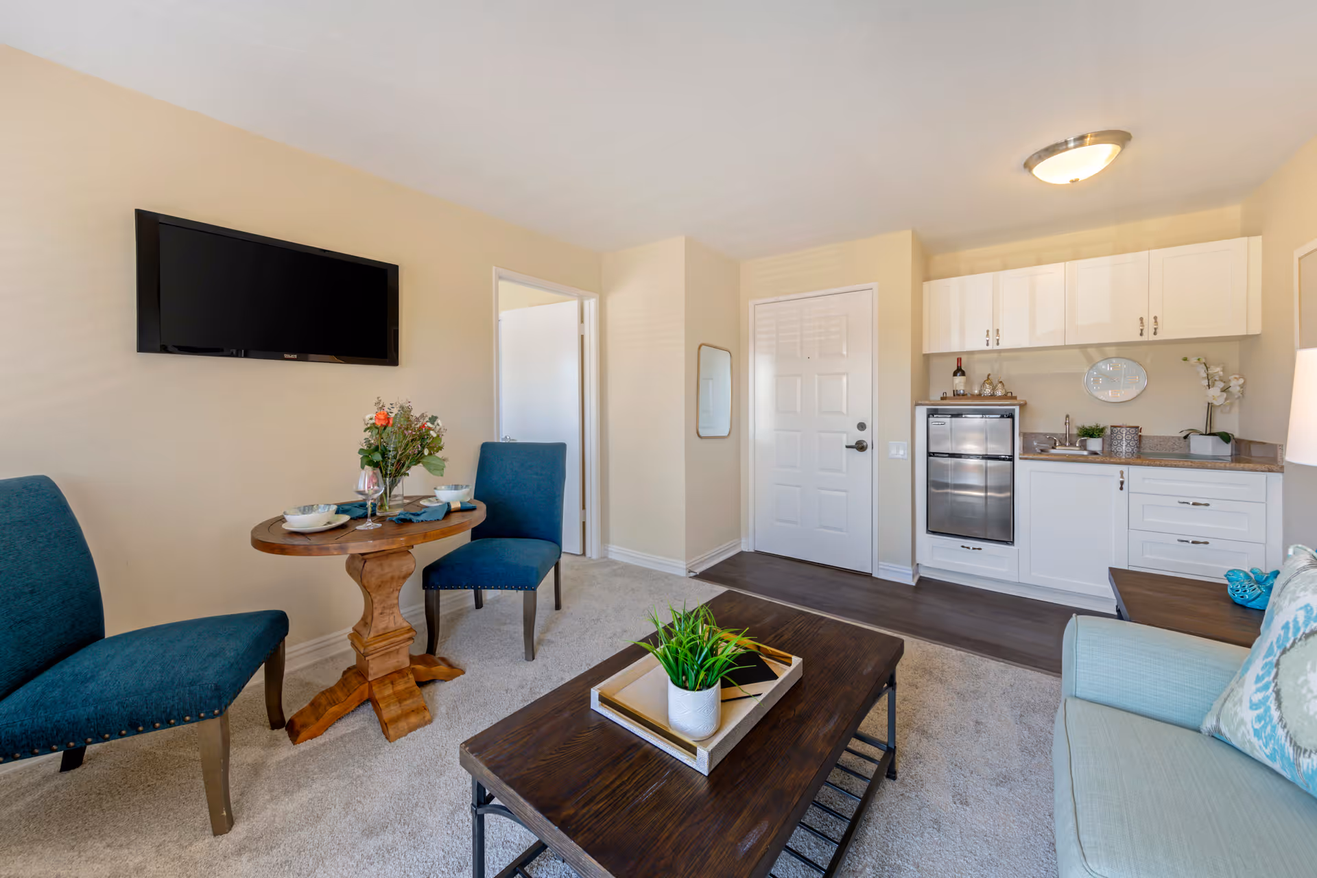 A cozy living area in a senior living facility featuring a mounted flat-screen TV on a beige wall, a small round wooden dining table set with two blue upholstered chairs, a coffee table with a plant centerpiece, and a kitchenette with white cabinets, a mini fridge, and countertop accessories. The room has a light carpet and a door leading to another room.