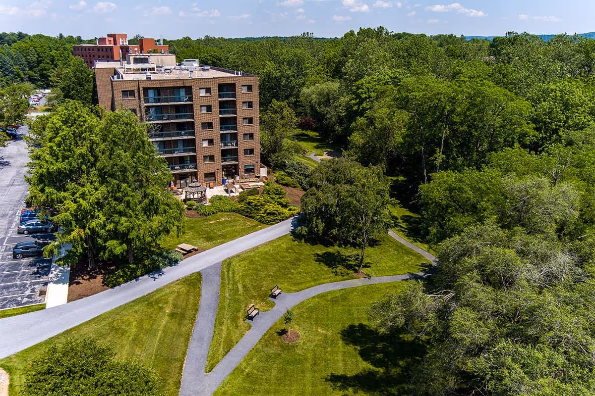 Aerial view of a multi-story brick senior living building with landscaped lawns, winding walking paths, benches, and surrounding trees.