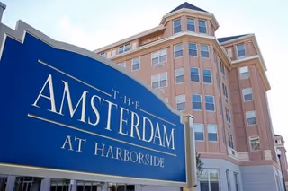 Exterior view of a multi-story residential building with a large blue sign in the foreground that reads 'The Amsterdam at Harborside'. The building has multiple windows and a light brown facade under a clear sky.