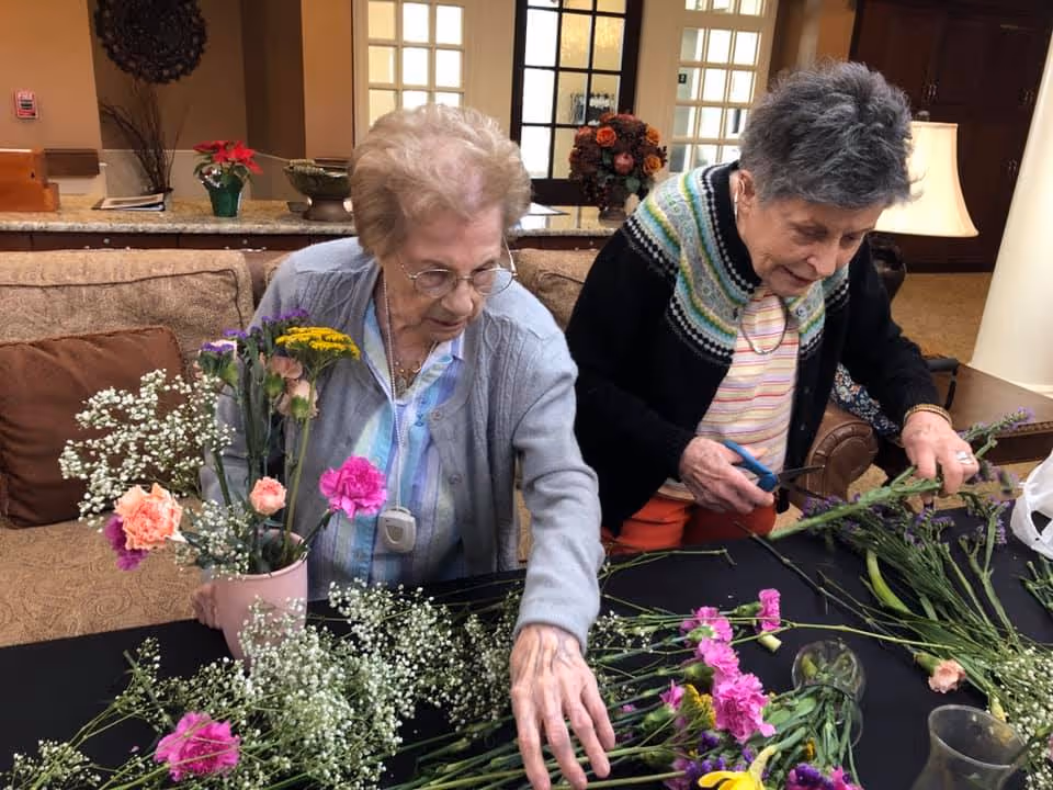 Two elderly women arranging various colorful flowers on a table in a cozy living room setting with a sofa and decorative items in the background.