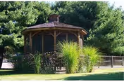 A wooden gazebo with a shingled roof surrounded by green grass, tall ornamental grasses, and pine trees in the background.