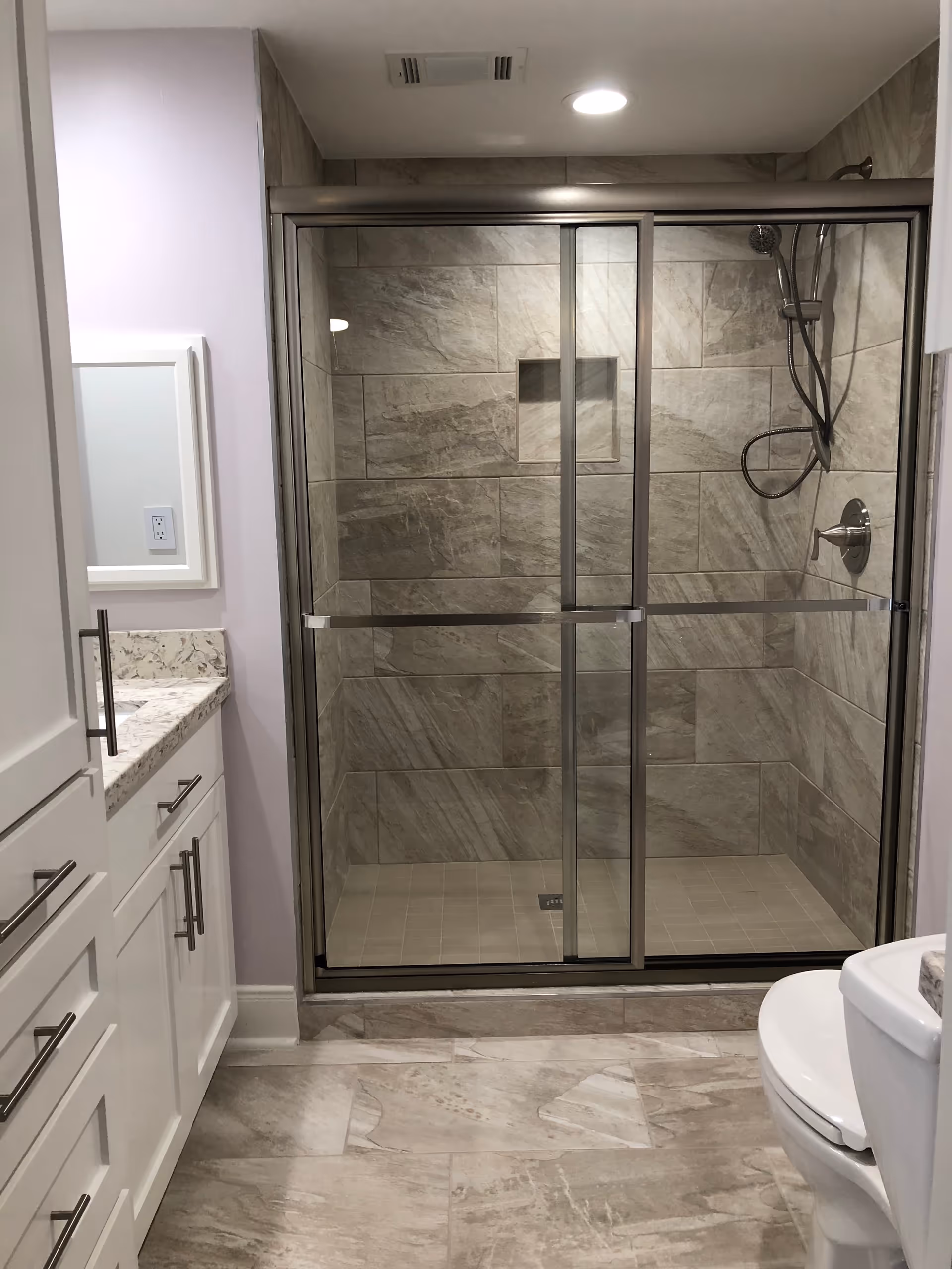 Modern bathroom featuring a glass-enclosed shower with beige and gray stone tiles, a white vanity with marble countertop and silver handles, and a white toilet.