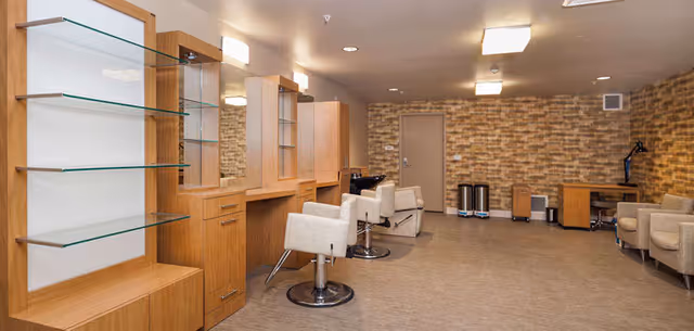 Interior view of a salon area with wooden cabinetry, glass shelves, mirrors, and two salon chairs. The room has a beige floor, brick-patterned wall, and additional seating with two armchairs and a desk with a lamp in the background.