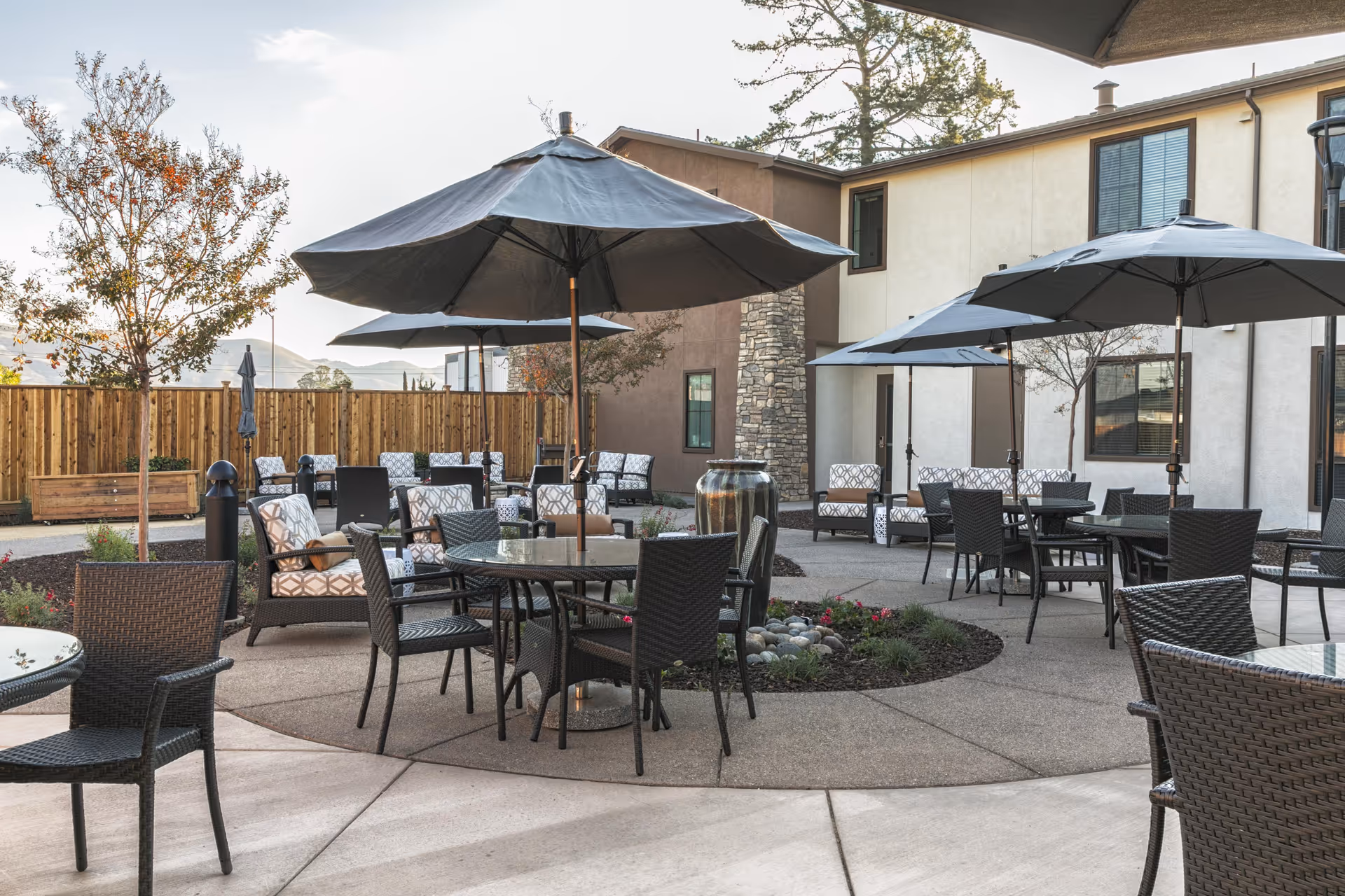 Outdoor courtyard patio with round tables, wicker chairs, and large umbrellas in front of a two-story building.