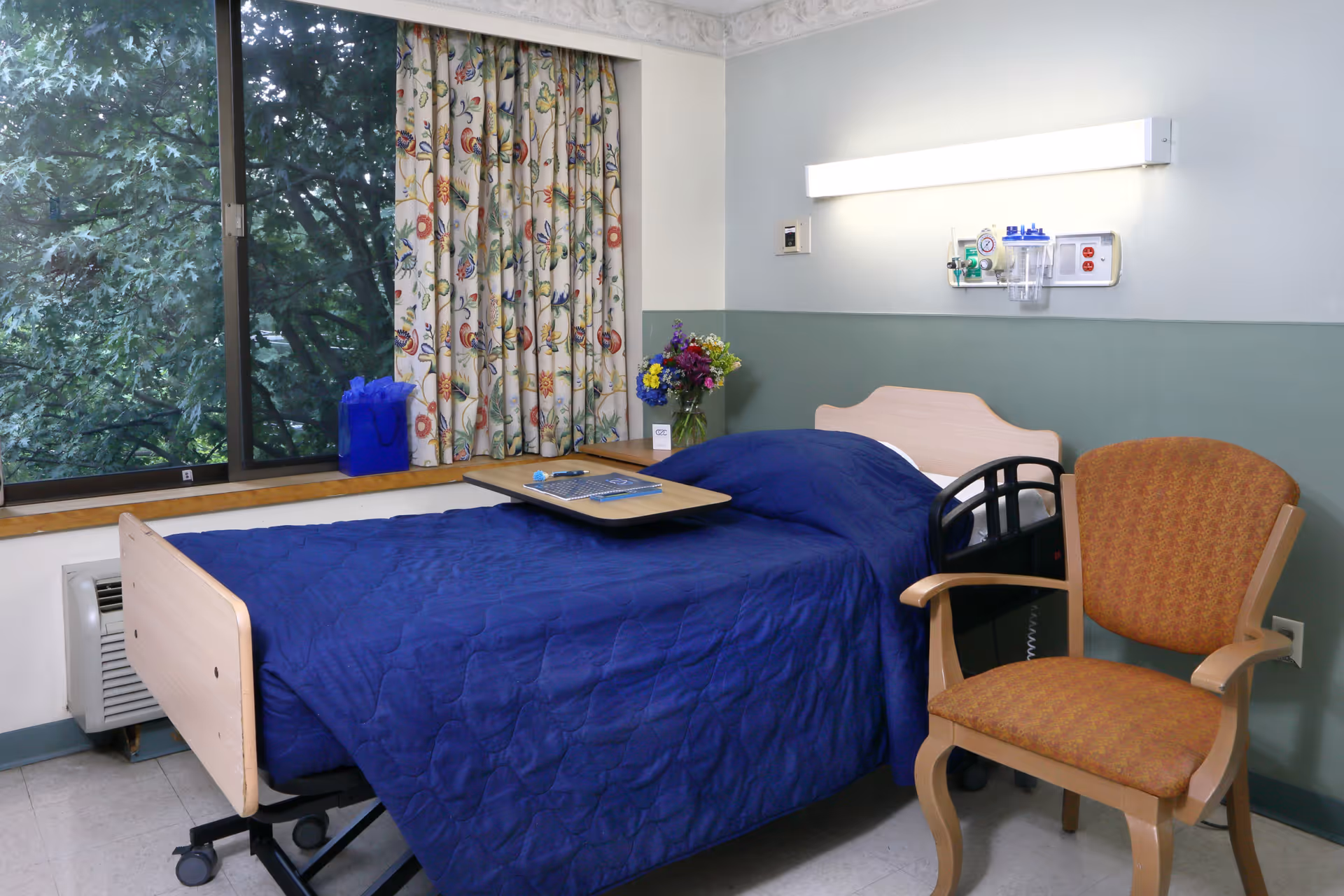 A patient room with a single hospital bed covered in a blue quilt, a small overbed tray, an armchair, and a window with floral curtains.