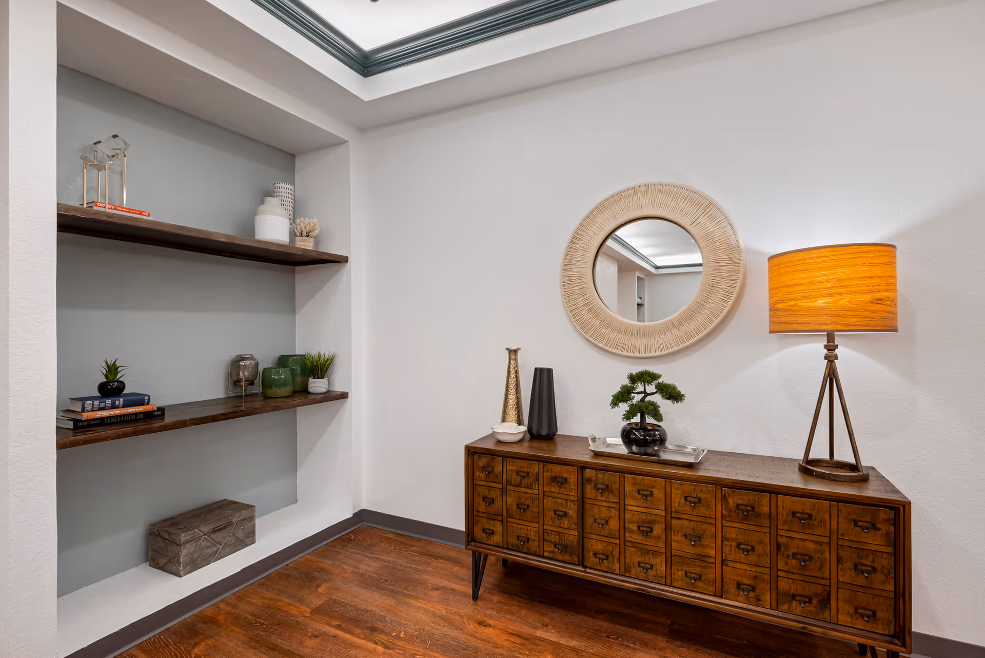 A cozy interior corner featuring a wooden sideboard with multiple small drawers, decorated with a bonsai plant, vases, and a tray. Above the sideboard hangs a round mirror with a textured frame. To the right, a tripod floor lamp with a warm orange shade provides soft lighting. On the left, two wooden shelves are mounted on a recessed wall, holding decorative items including books, small plants, and vases. The room has wooden flooring and white walls with a gray accent in the recessed area.