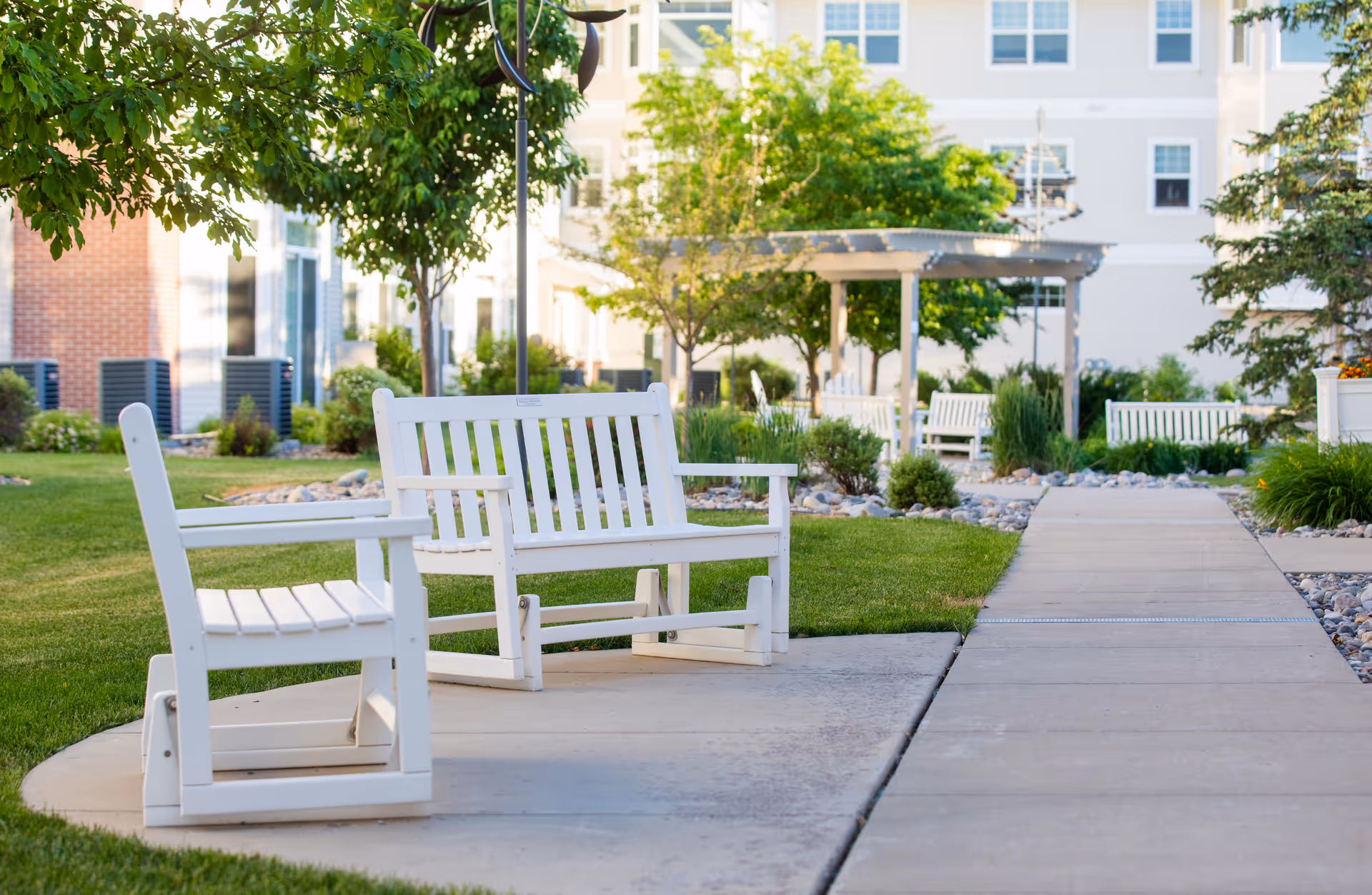 Outdoor seating area with white wooden benches and chairs on a paved pathway surrounded by green grass, trees, and shrubs in front of a multi-story building.