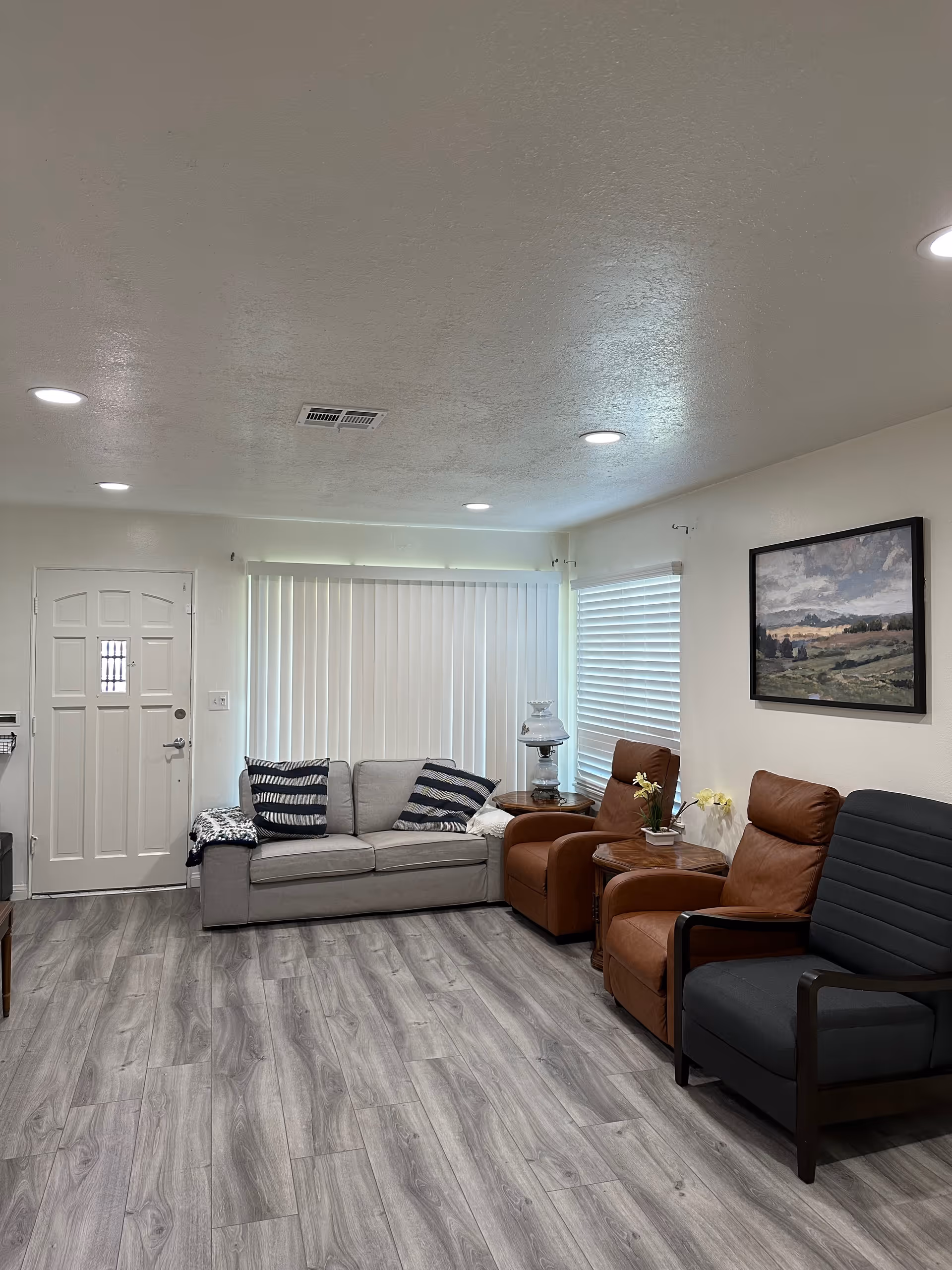 Bright living room with a sofa, recliner chairs, side tables, wall art, and a front door with vertical blinds.